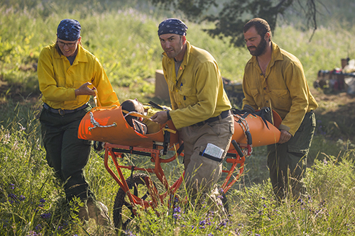 Three wildland firefighters use a wheeled liter to transport an injured person up a grassy hill