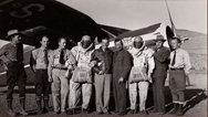 1935 photo of nine men in the Aerial Fire Control Experimental Project standing in front of plane, two in parachute gear.