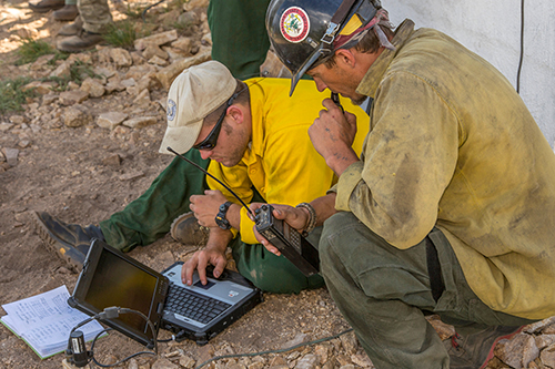 A fire fighter squats next to a division supervisor who is sitting on the ground reviewing data on a laptop sitting in front of him. Decorative.