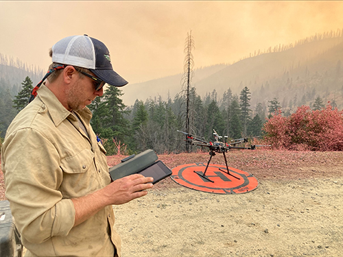 Unmanned aircraft technician using a tablet to operate a drone that sits in background on launch pad. Decorative