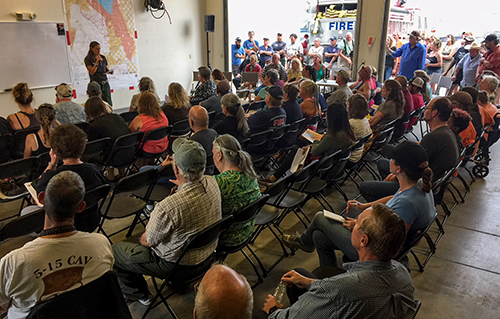 A large group of people seated in rows inside a fire house garage, listen to a briefing being given by one person at the front. People also standing outside at the open doors listening. .  Decorative.
