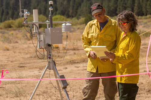 Two fire weather specialist standing in open area in front of a RAWS weather station reading documents.  Decorative.