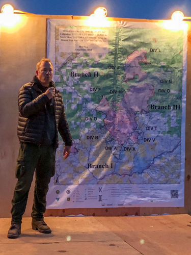 a wildland fire incident commander stands on a plywood podium in front of a large map talking on a microphone. Decorative