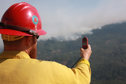 Photo of A wildland firefighter holds a weather meter out in front of him taking readings of the terrain. Decorative.