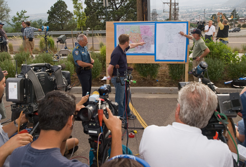 Two people pointing at large maps on a board outdoors, while news crews record the briefing. .  Decorative