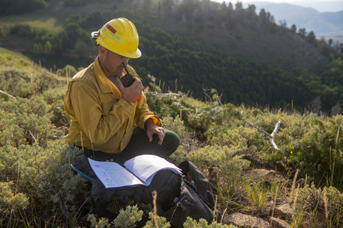 Photo of A wildland firefighter crouches on a mountain ridge while talking into a hand radio. Decorative.