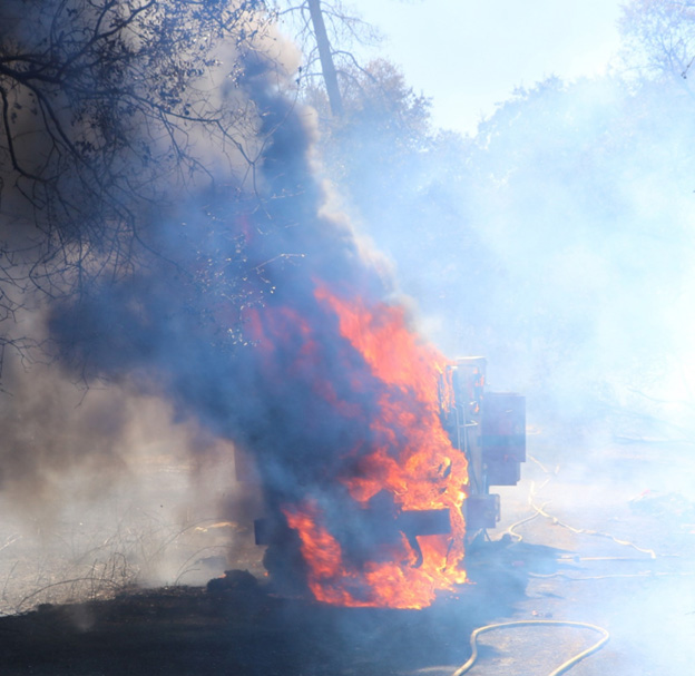 In a forested area, a fire engine in flames, heavy smoke surrounding the engine. 