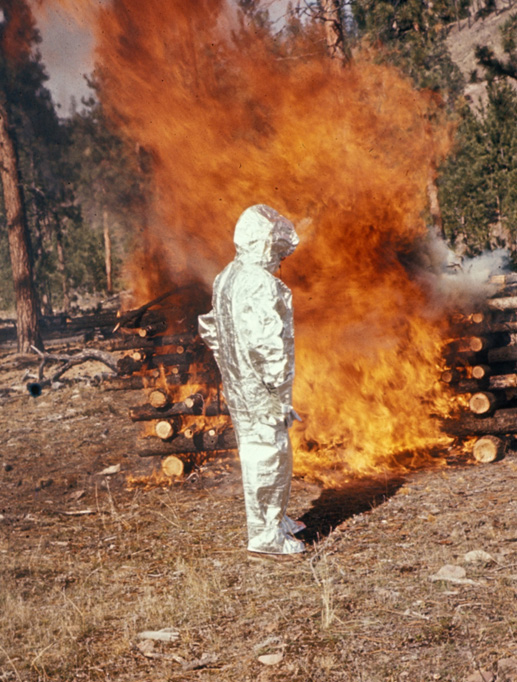 Main in a testing suit standing before a contained fire in the forest.