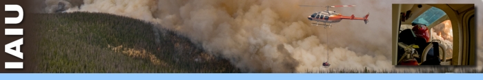 IAIU header graphic.  A helicopter flying over dense smoke from burning timber below. An aerial ignition tank is suspended from the helicopter. An inset photo of a helicopter pilot looking out window to wildland fire below. Decorative.