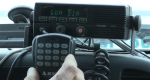 Photo of hand holding keypad to a mobile radio mounted on the dash board of a vehicle.