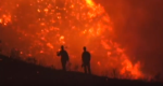 Photo of silhouette of fire fighters walking up hill with flames in background.