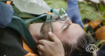 Man laying on ground with oxygen mask on and a hand holding his head.