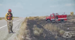Firefighter walking on side of road looking at a burned out area with a fire truck sitting in burned out area in the distance.
